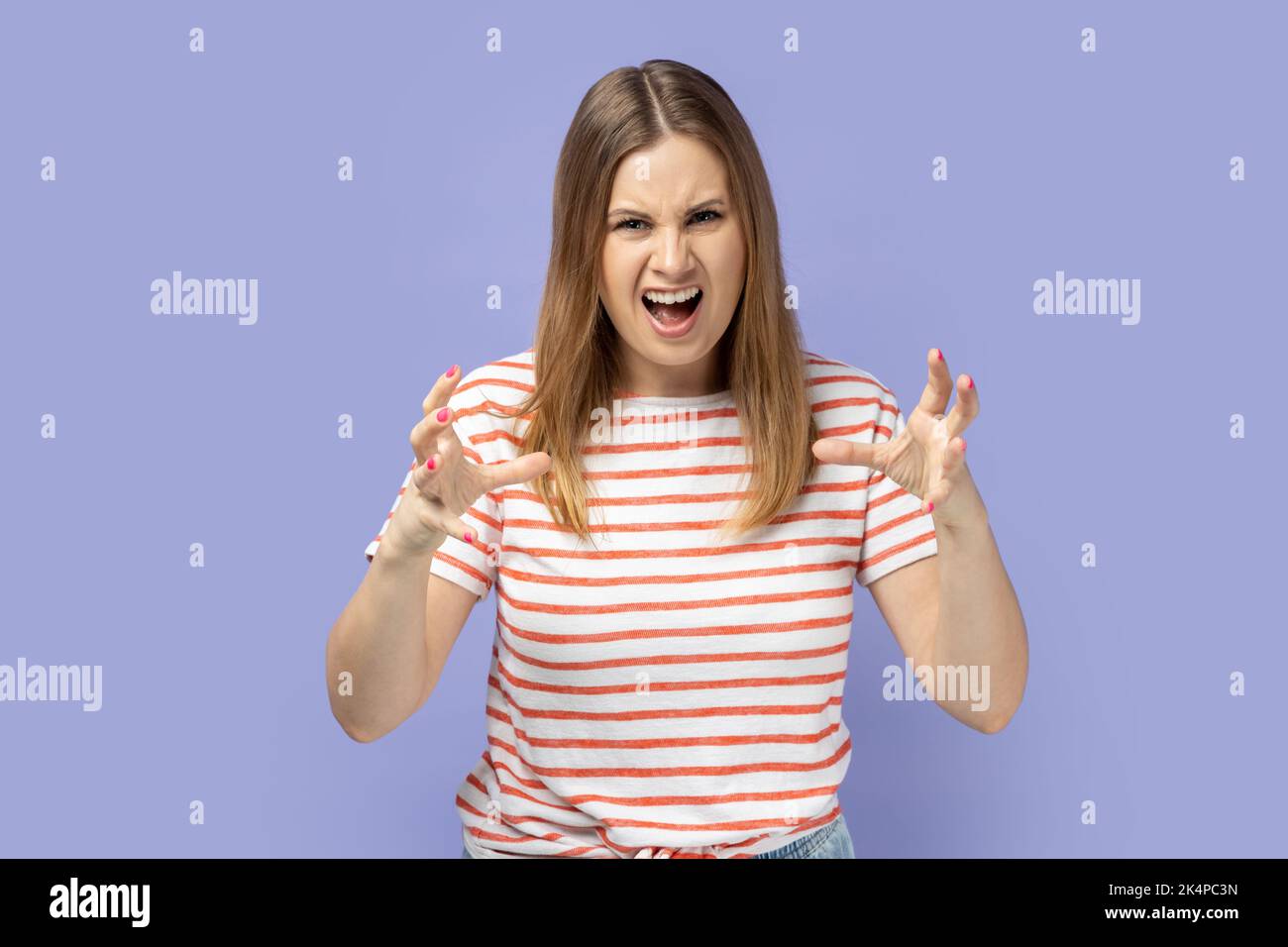 Porträt einer wütenden jungen blonden Frau mit gestreiftem T-Shirt, das aggressive Emotionen ausdrückt, die erhobenen Arme hebt und vor Hass und Wut schreit. Innenaufnahme des Studios isoliert auf violettem Hintergrund. Stockfoto
