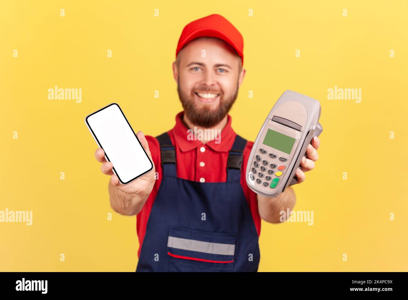 Porträt eines zufriedenen bärtigen Arbeiters, der POS-Terminal und Smartphone mit leerem Display für Werbetexte stehend und aushaltend. Innenaufnahme des Studios isoliert auf gelbem Hintergrund. Stockfoto