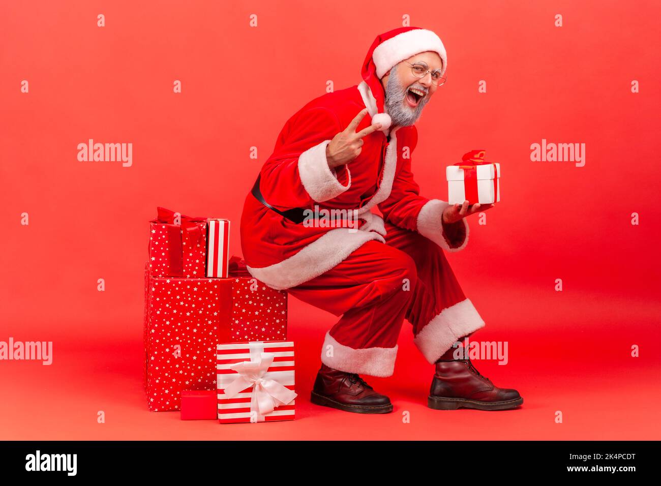 Positiv aufgeregt älterer Mann mit grauem Bart im weihnachtsmann Kostüm sitzt auf großer Box mit Weihnachtsgeschenk, hält Geschenk, zeigt V-Zeichen zur Kamera. Innenaufnahme des Studios isoliert auf rotem Hintergrund. Stockfoto