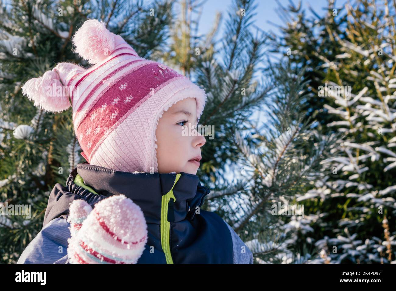 Kleines Mädchen 3-4 Jahre alt in Winterkleidung Overalls, Hut und Fäustlinge, steht im Freien gegen schneebedeckte Kiefern und Tannen und schaut weg Stockfoto