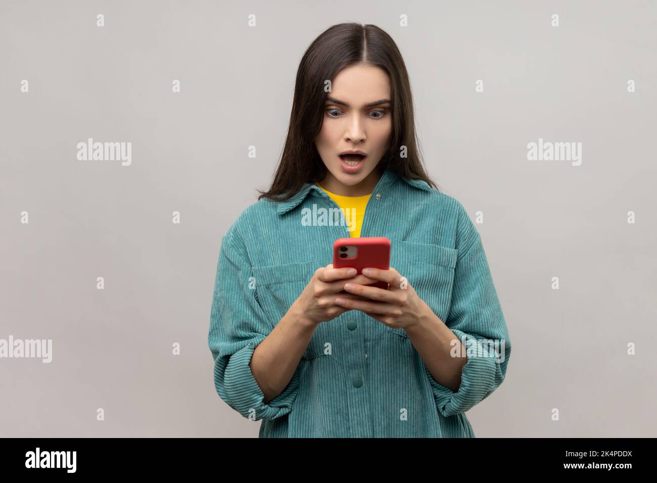 Schockierte hübsche Frau, die ihre Nachricht auf dem Smartphone mit offenem Mund liest, mobile Geräte für die Kommunikation nutzt, im Internet surft und eine Jacke im lässigen Stil trägt. Innenaufnahme des Studios isoliert auf grauem Hintergrund. Stockfoto
