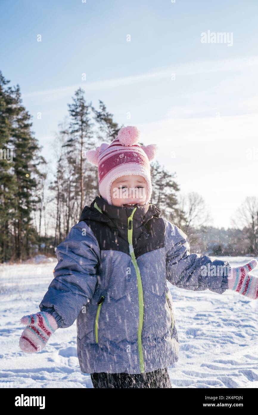 Kleines Mädchen, 3-4 Jahre alt, im Winter Overalls, Hut und Fäustlinge, steht im Freien gegen blauen Himmel und Tannen und wirft Schnee über sie Stockfoto