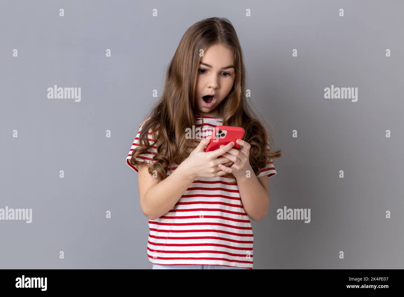 Porträt eines staunenden kleinen Mädchens, das gestreiftes T-Shirt mit Smartphone trägt, auf den Bildschirm des Geräts schaut und Post für den Blog ihres Kindes schreibt. Innenaufnahme des Studios isoliert auf grauem Hintergrund. Stockfoto