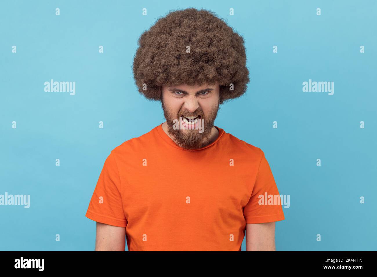 Porträt eines wütenden Mannes mit Afro-Frisur, das ein orangefarbenes T-Shirt trägt und die Kamera anschaut, mit aggressivem Gesichtsausdruck, der negativ, Hass ausdrückt. Innenaufnahme des Studios isoliert auf blauem Hintergrund. Stockfoto