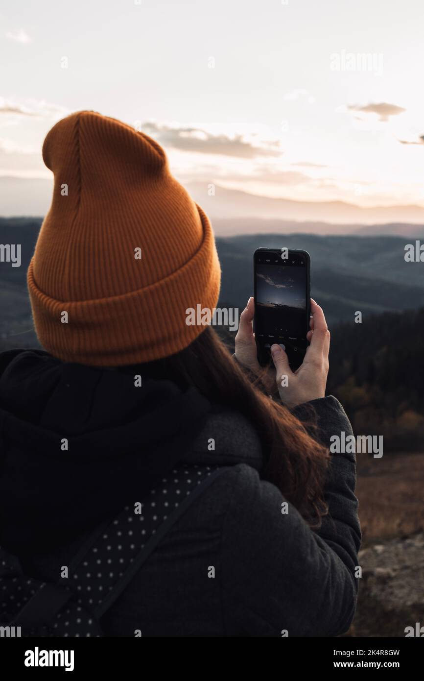 Eine junge Frau mit orangefarbener Beanie macht ein Foto vom Sonnenuntergang in den Bergen Stockfoto