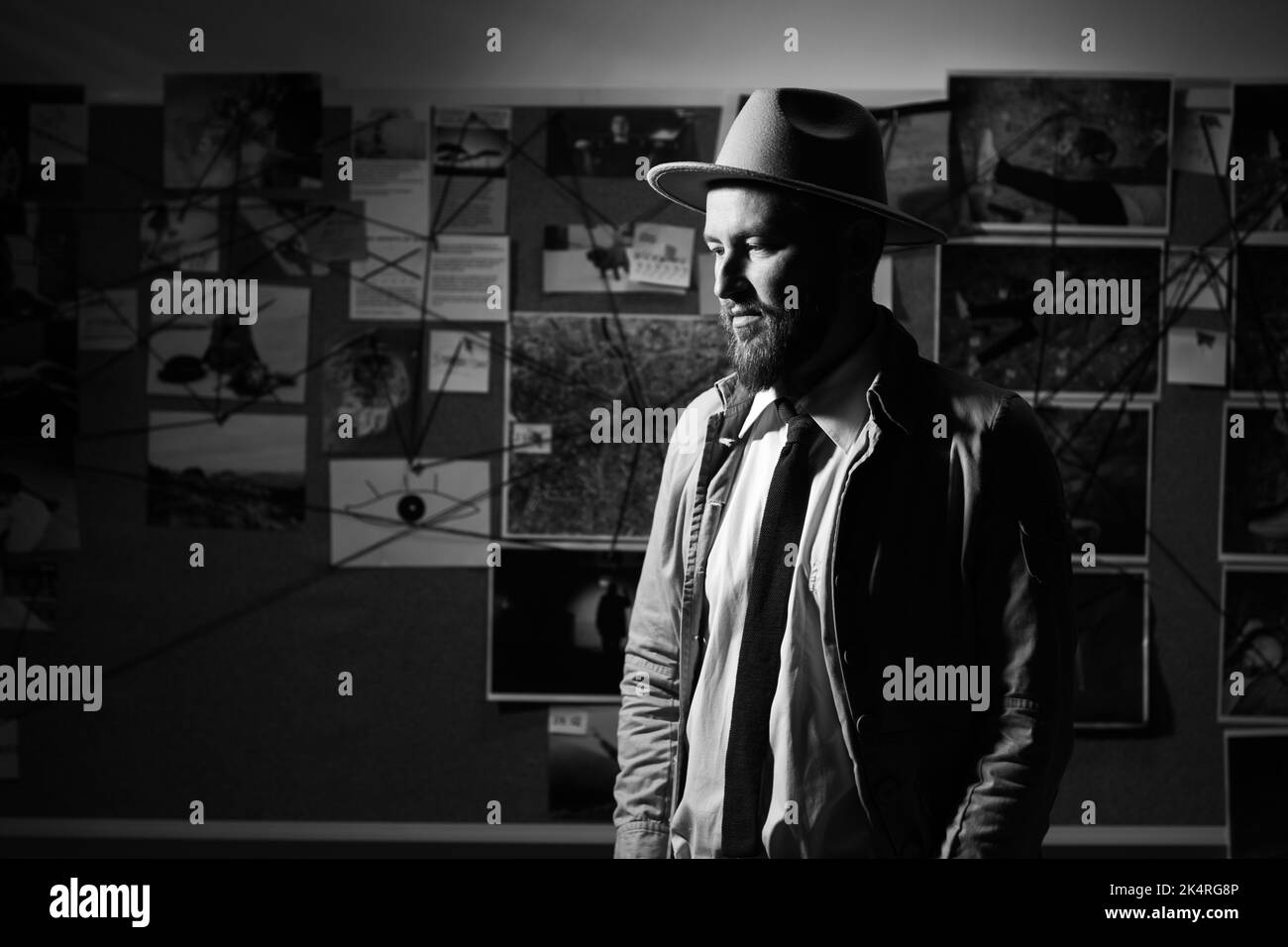 Junger Spionageagent mit Hut, Trenchcoat und Krawatte, Detektiv bei der Arbeit im Büro spät in der Nacht, Hintergrund der Beweistafel, schwarz-weiß-Schuss Stockfoto