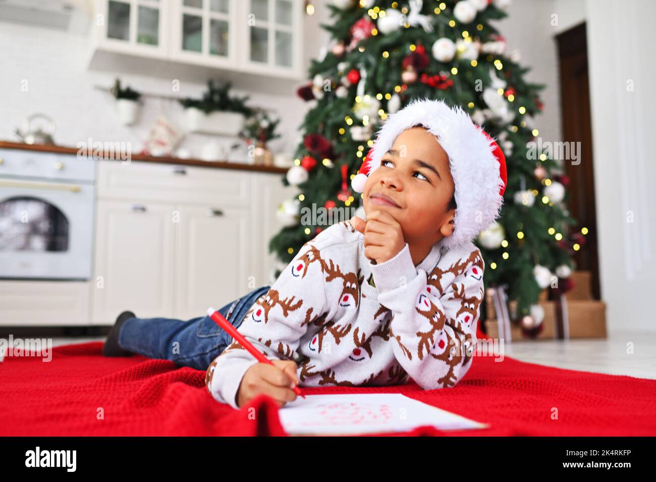 Der Junge im weihnachtsmütze, der auf einem karierten Tuch liegt, lächelt träumerisch und schreibt einen Brief an den Weihnachtsmann auf ein weißes Blatt - eine Wunschliste auf dem Hintergrund eines Weihnachtsfestes Stockfoto
