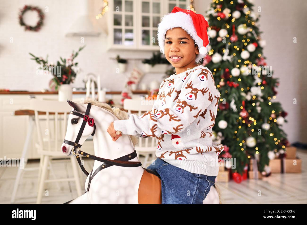 Der kleine afroamerikanische Junge in einem grauen Weihnachtspullover und einem roten weihnachtsmann-Hut lächelt, während er auf einem schaukelnden Spielzeug-Pferd vor dem Hintergrund eines sitzt Stockfoto