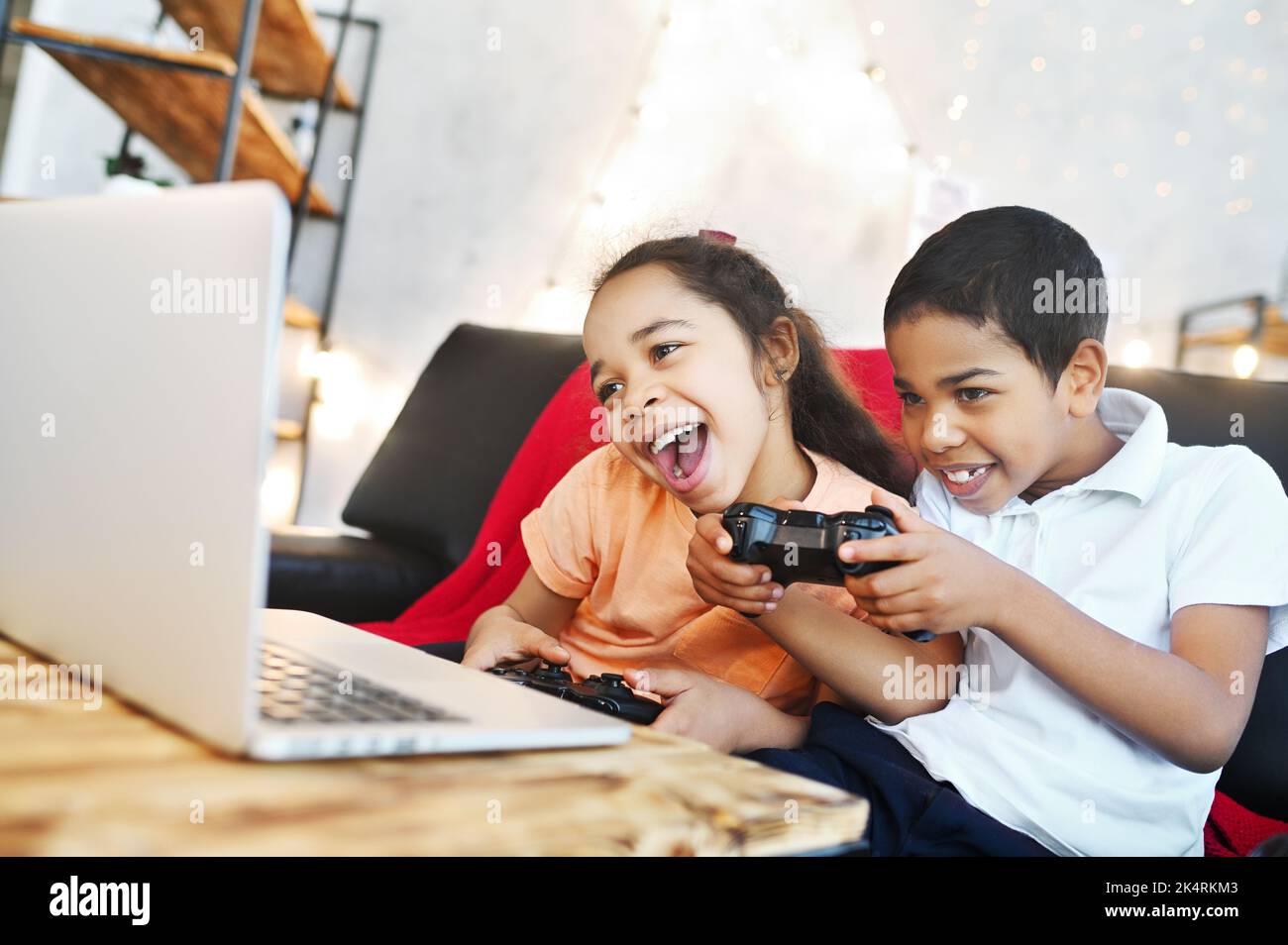 Afroamerikanische Kinder - ein Junge und ein Mädchen halten ein Gamepad in den Händen, spielen Computerspiele auf einem Laptop und lachen, während sie auf der Couch sitzen. Stockfoto