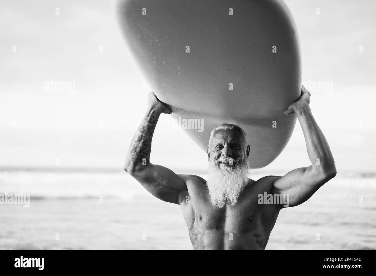 Glücklicher Senior Surfer Mann hält Surfbrett am Strand bei Sonnenuntergang - Fokus auf Gesicht - Schwarz-Weiß-Schnitt. Stockfoto