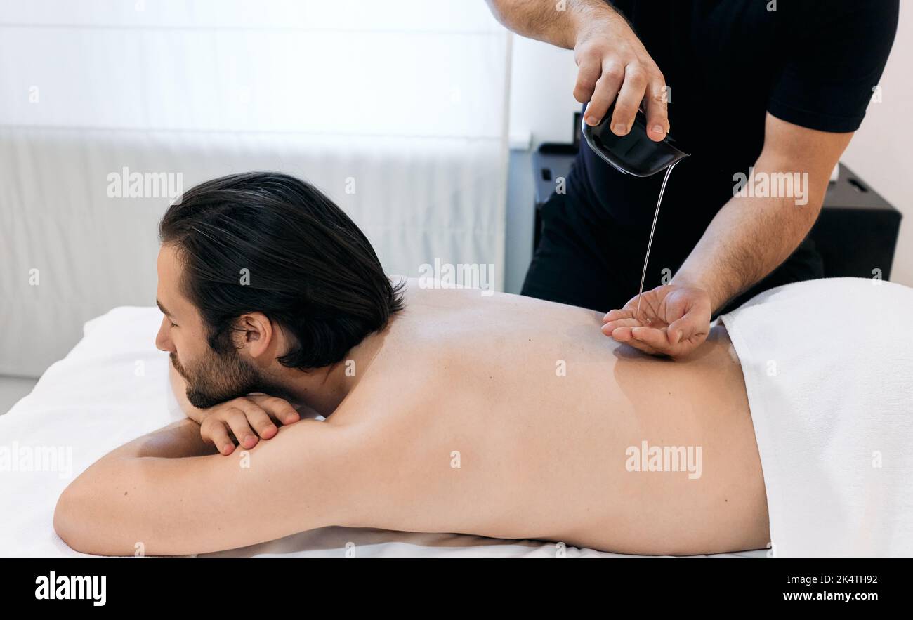 Masseur wendet Aromaöl auf den Körper des erwachsenen Mannes während der Massage und Aromatherapie im Massagesalon an Stockfoto