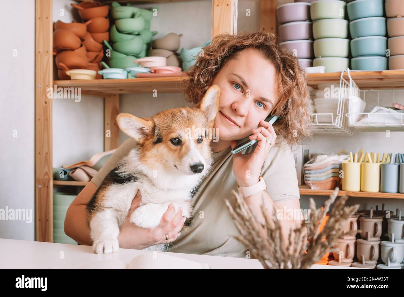 Lächelnd konzentrierte Geschäftsfrau spricht Smartphone, hält niedlichen ruhigen Hund Corgi in der Werkstatt. Set aus Silikon-Babyschalen in der Nähe der Wand auf Regalen. Zu Hause Stockfoto