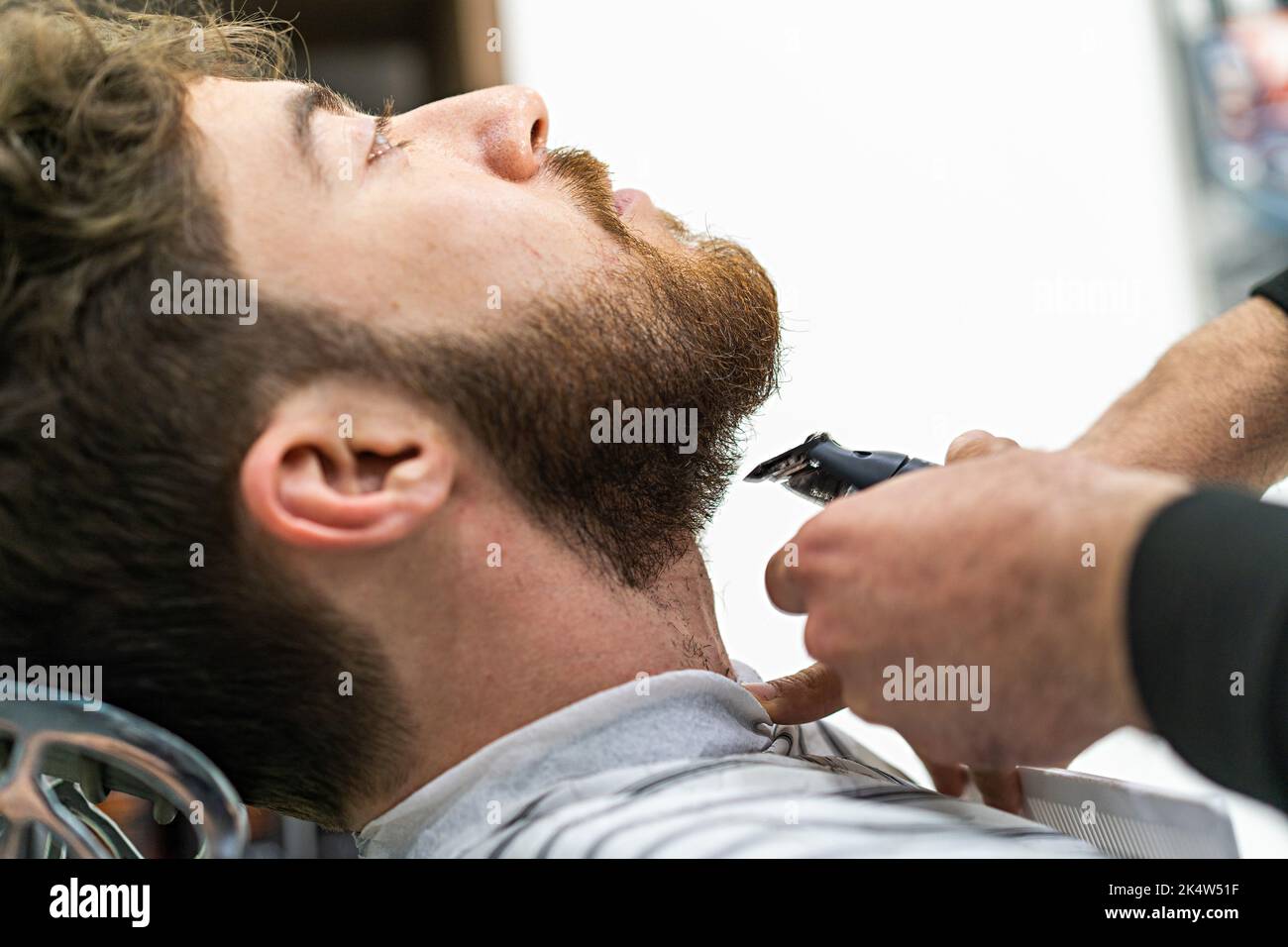 Ein bärtiger Kunde eines Rasiersalons hat seinen Bart mit einem Haarschneider getrimmt - Detail mit der Hand des Barbiers auf dem Hals des Mannes, der auf dem Armchai sitzt Stockfoto