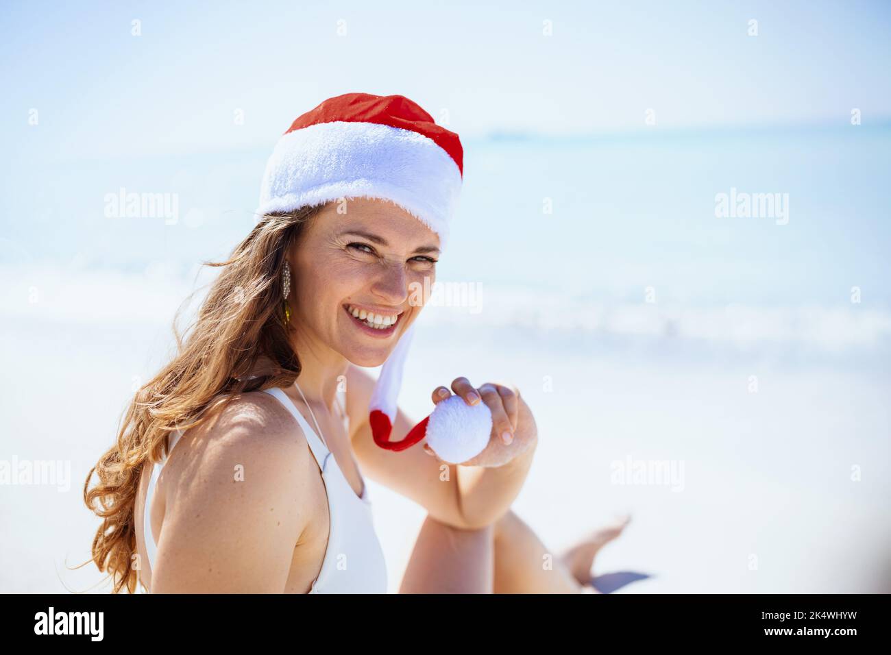 Fröhliche, stilvolle Frau im weißen Badeanzug mit gestreiftem weihnachtshut am Strand. Stockfoto