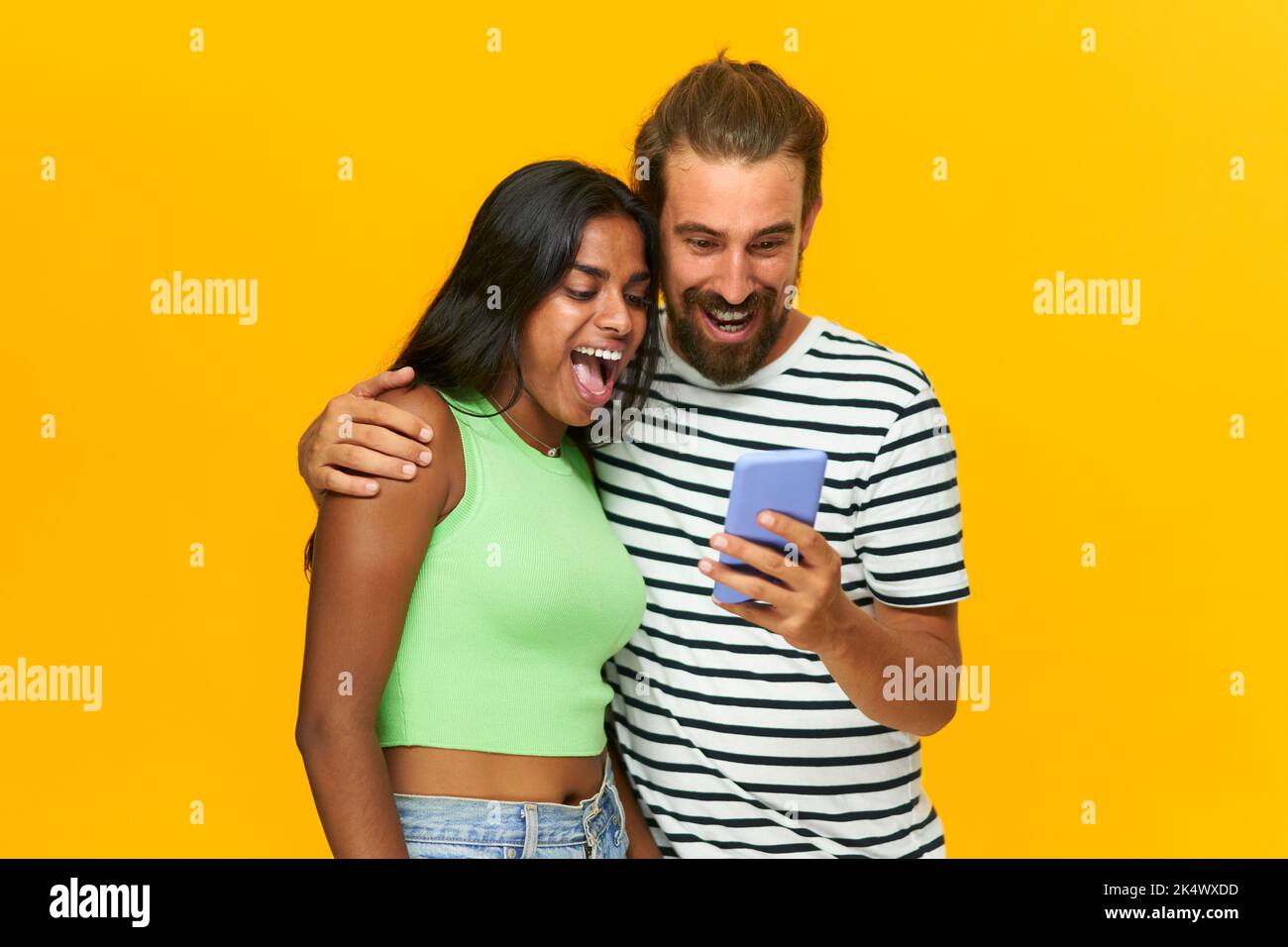 Glückliches multirassisches Paar überrascht Blick auf ein Mobiltelefon, isoliert über gelbem Hintergrund. Stockfoto