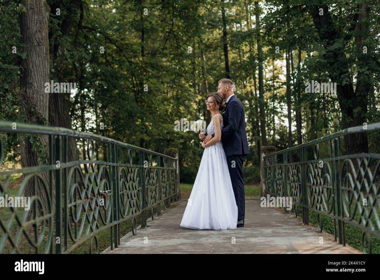 Im Sommer steht ein tolles Hochzeitspaar im Park. Junge hübsche Frau mit Blumenstrauß, Bräutigam in Anzug umarmt Braut. Stockfoto