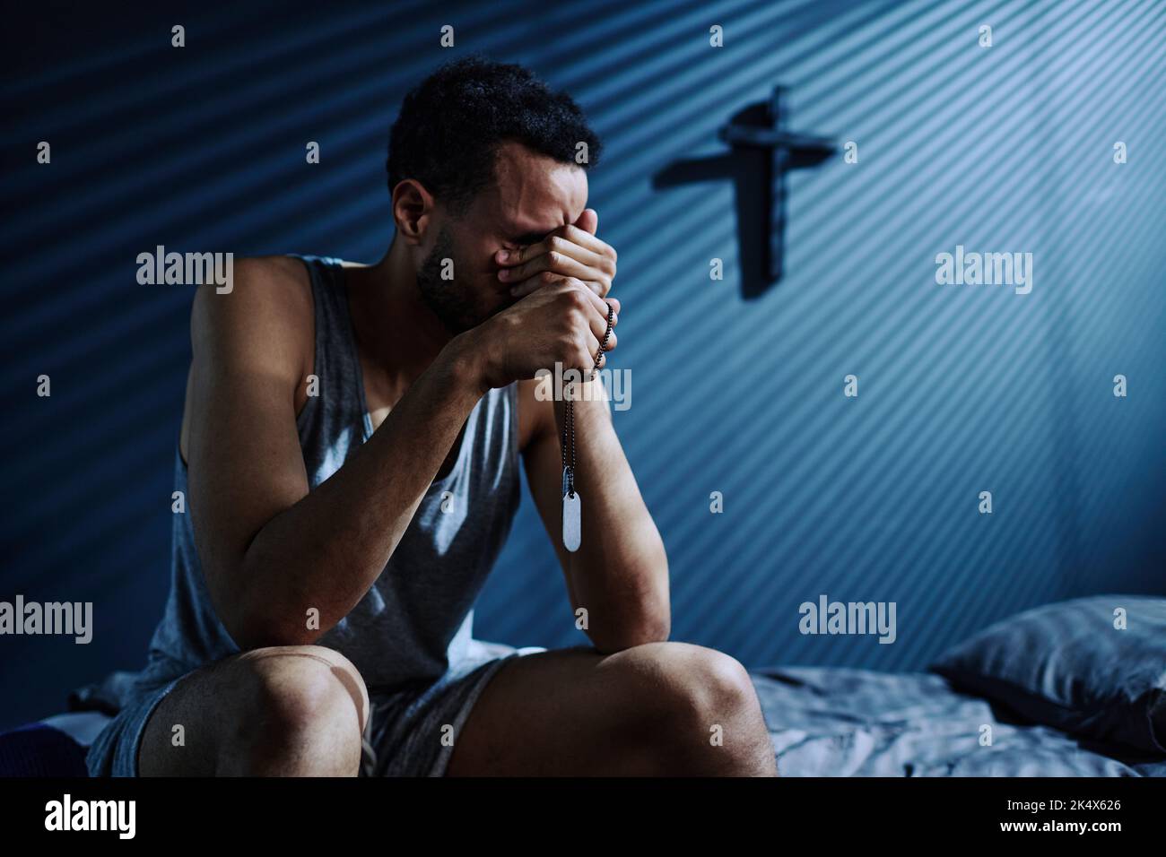 Junger depressiver Mann mit zwei Medaillons auf silberner Kette weint oder trauert über den Verlust von jemandem, den er liebte, während er nachts auf dem Bett saß Stockfoto