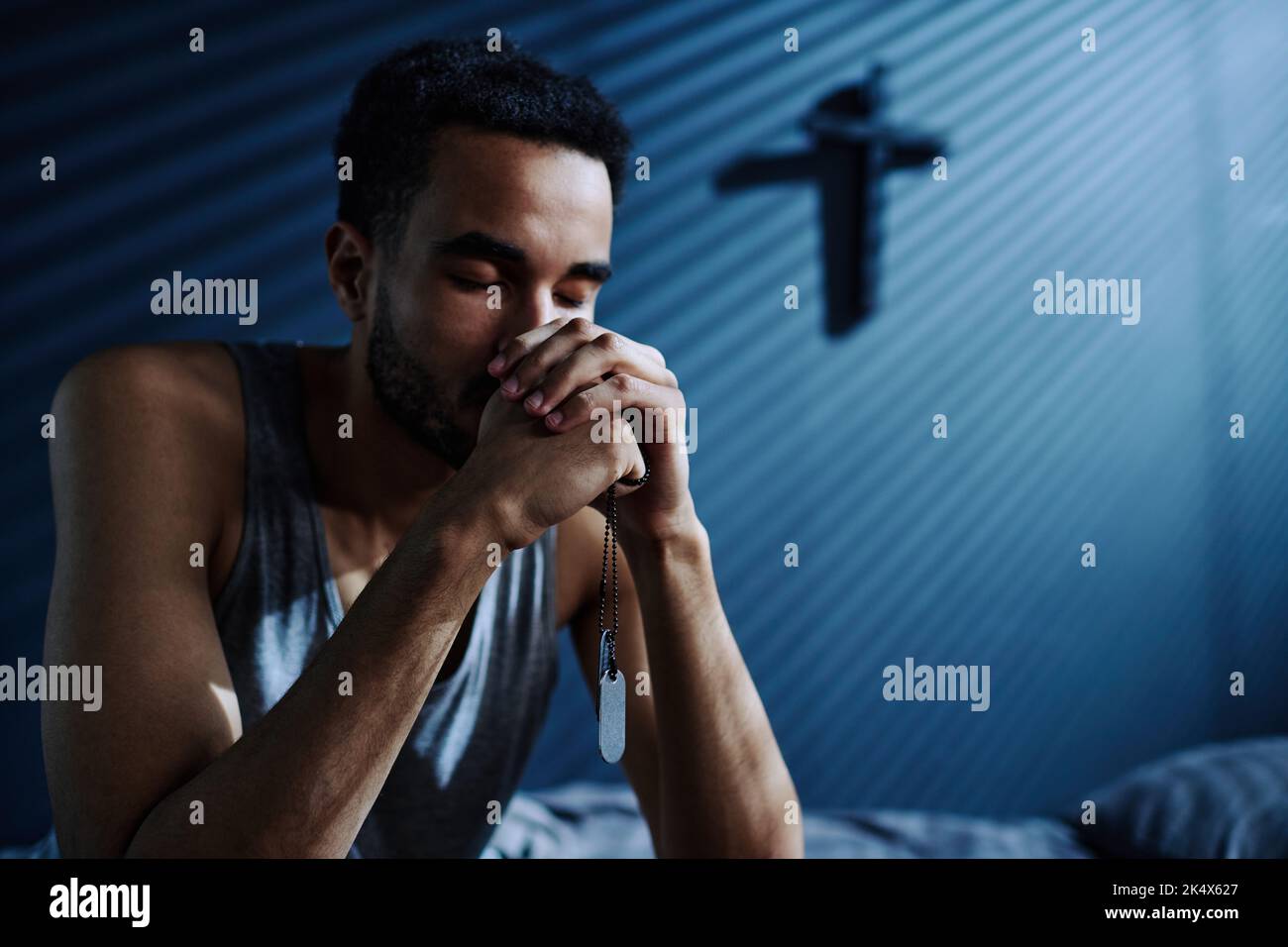 Junger depressiver Mann mit Metallmedaillons an der Kette in den Händen, der Gott bittet, ihm beim Schlafen zu helfen, während er auf dem Bett sitzt Stockfoto
