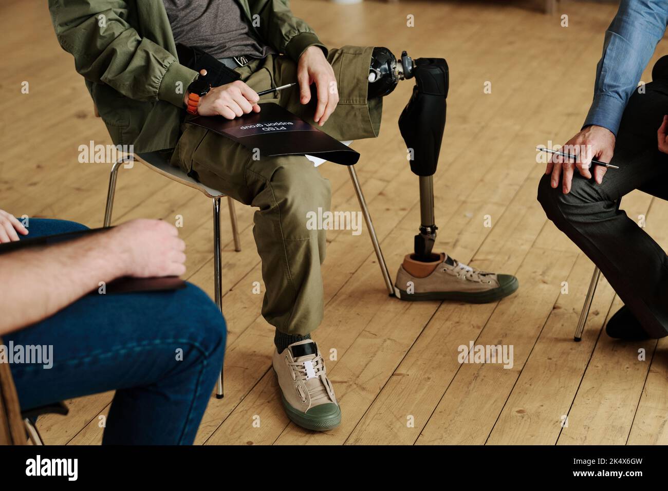 Nahaufnahme der Beine eines jungen Mannes mit körperlicher Behinderung, der unter anderen Männern auf einem Stuhl sitzt, während er über ihre psychischen Probleme diskutiert Stockfoto