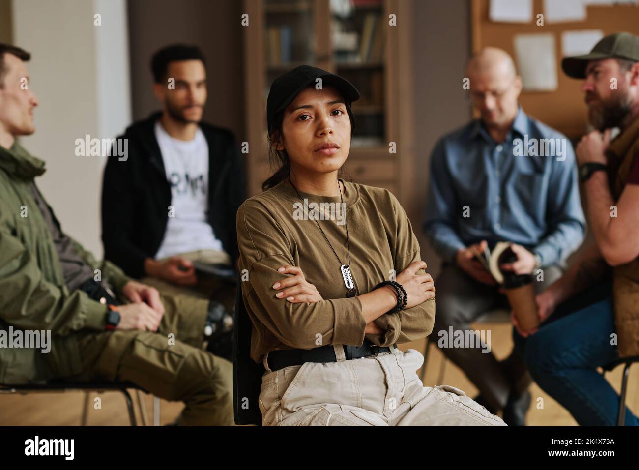 Junge gestresste hispanische Frau mit gekreuzten Armen auf der Brust und Blick auf die Kamera, während sie gegen interkulturelle Männer sitzt, die diskutieren Stockfoto