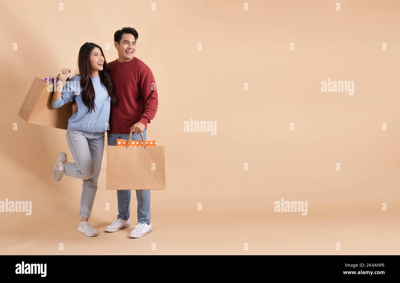 Glücklich lächelndes asiatisches Paar mit Einkaufstaschen im Pullover genießen ihren Winterrabatt Verkauf isoliert auf beigefarbenem Hintergrund. Shopaholic modische 20s Mann und Frau, die weg mit Kopieplatz schauen. Stockfoto