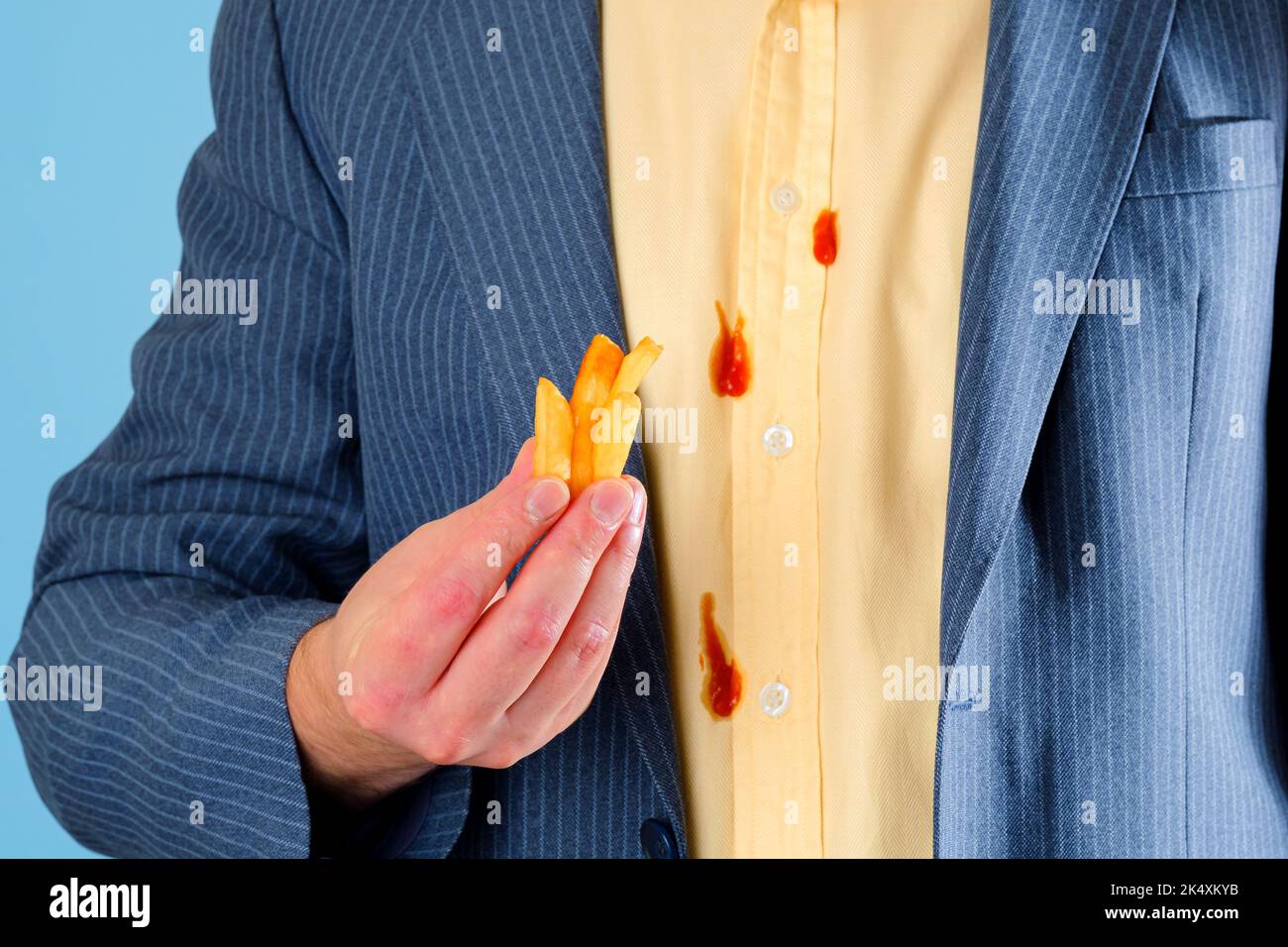 Pommes Frites in der Hand eines Mannes vor dem Hintergrund von Ketchup-Flecken auf einem gelben Hemd. Isoliert auf blauem Hintergrund Stockfoto