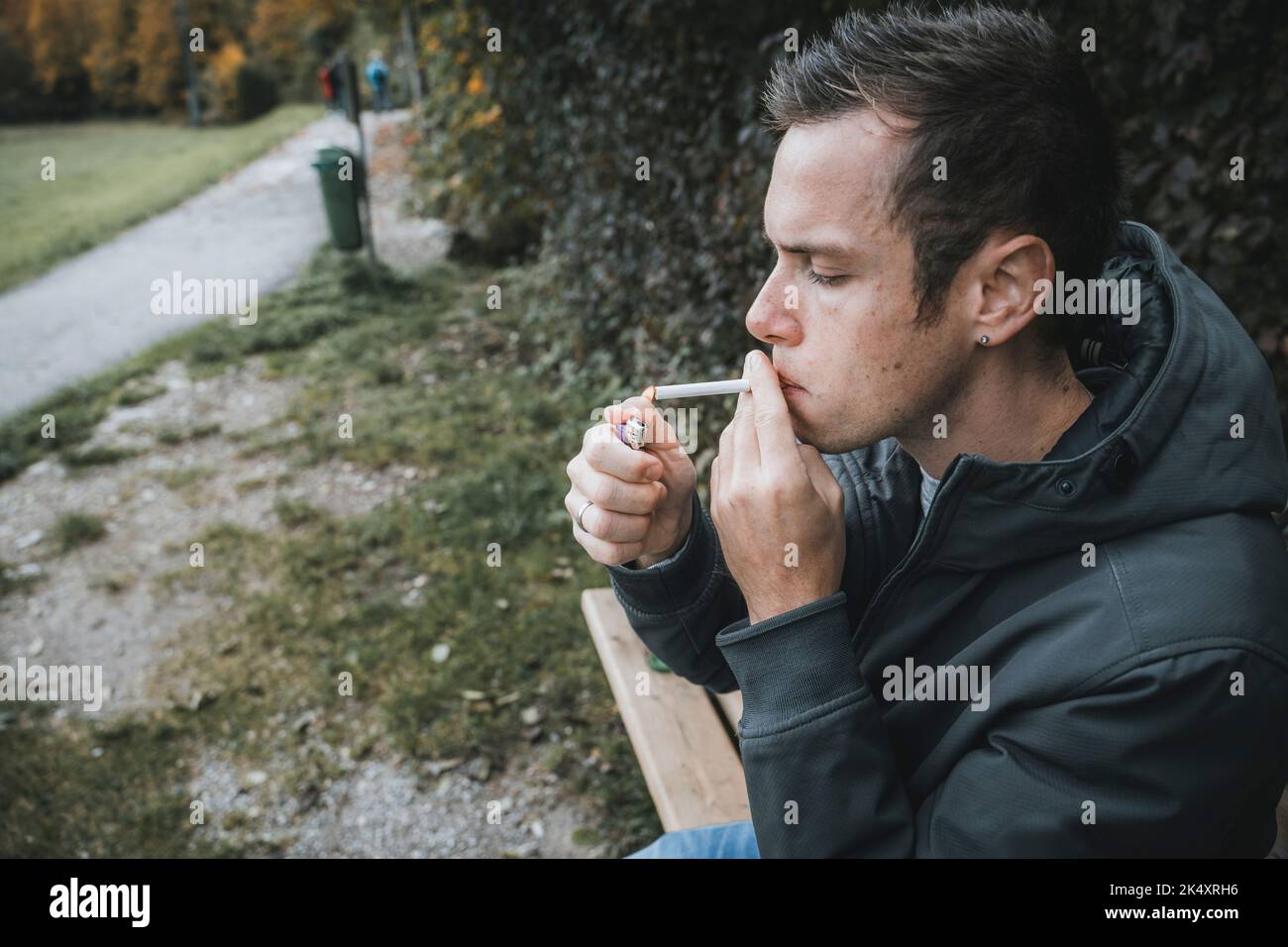 03. Oktober 2022: Junger Mann sitzt auf Einer Parkbank und raucht Eine Zigarette. Smoke zündet Eine Zigarette mit Feuerzeug an Stockfoto
