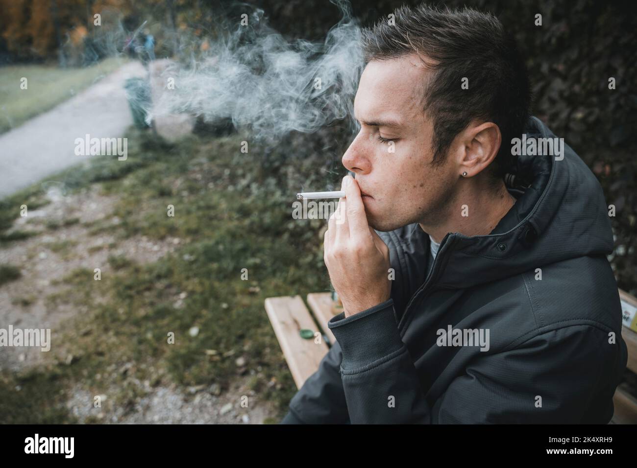 03. Oktober 2022: Junger Mann sitzt auf Einer Parkbank und raucht Eine Zigarette. Raucher zieht eine brennende Zigarette, Symbol Bild Rauchen Stockfoto