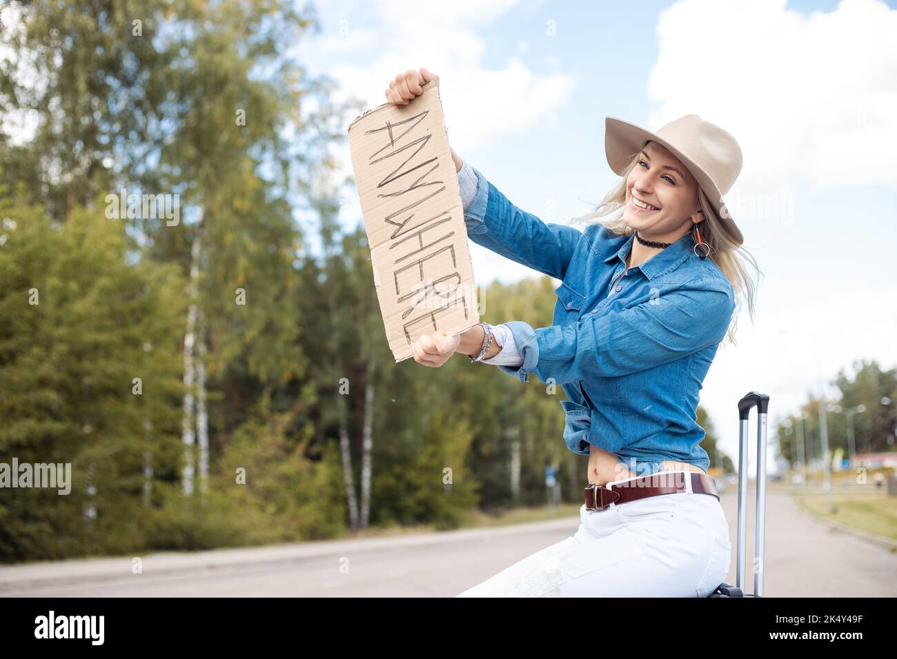 Junge erstaunliche blonde Frau, die Hände mit Pappe mit Inschrift überall, sitzt auf schwarzem Koffer in der Nähe des Waldes. Stockfoto