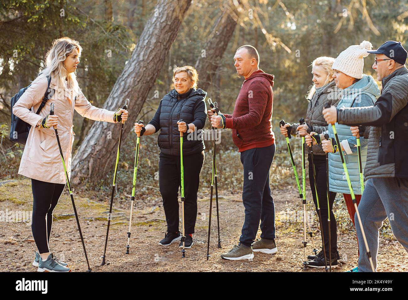 Blonde Trainerin, Sporttrainerin mit einer Gruppe von Menschen lehren skandinavisches Gehen im Wald. Leitender Körperkurs Stockfoto