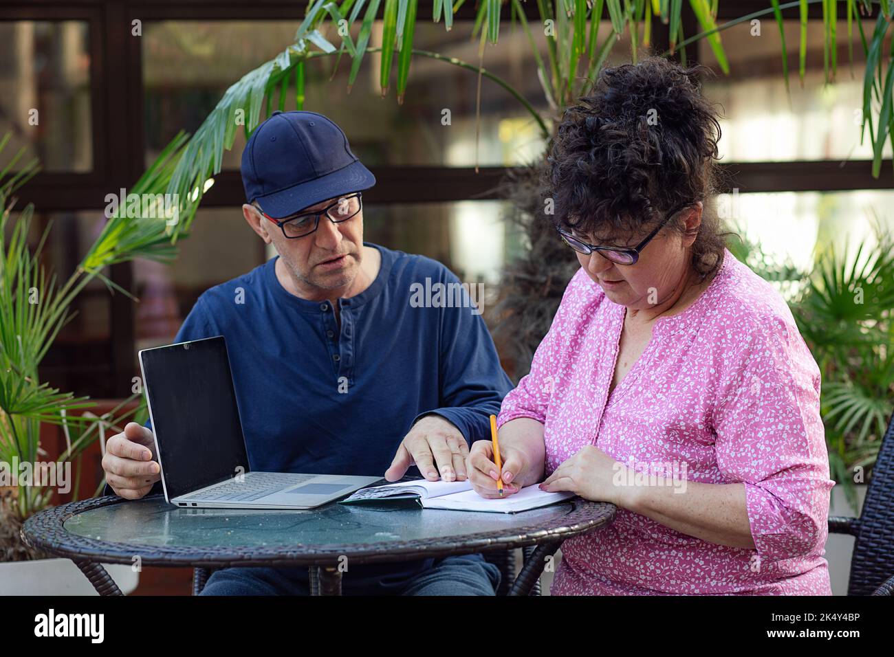 Reifer Mann und Frau in einer Brille reden miteinander, sitzen am Tisch, schreiben auf einem Notizblock neben dem Laptop unter der Palme Stockfoto