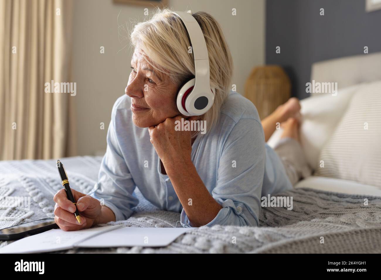 Glückliche ältere kaukasische Frau, die im Schlafzimmer auf dem Bett lag, Notizen gemacht und Kopfhörer getragen hat Stockfoto