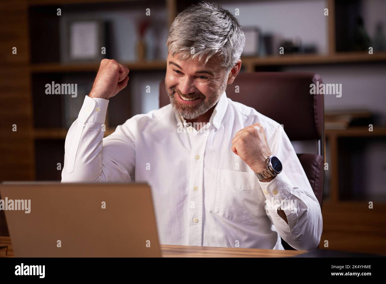 Der glückliche Geschäftsmann war von den guten Nachrichten überrascht und machte eine Ja-Geste, während er auf den Laptop im Büro schaute. Trader genießen Erfolg, feiern Stockfoto