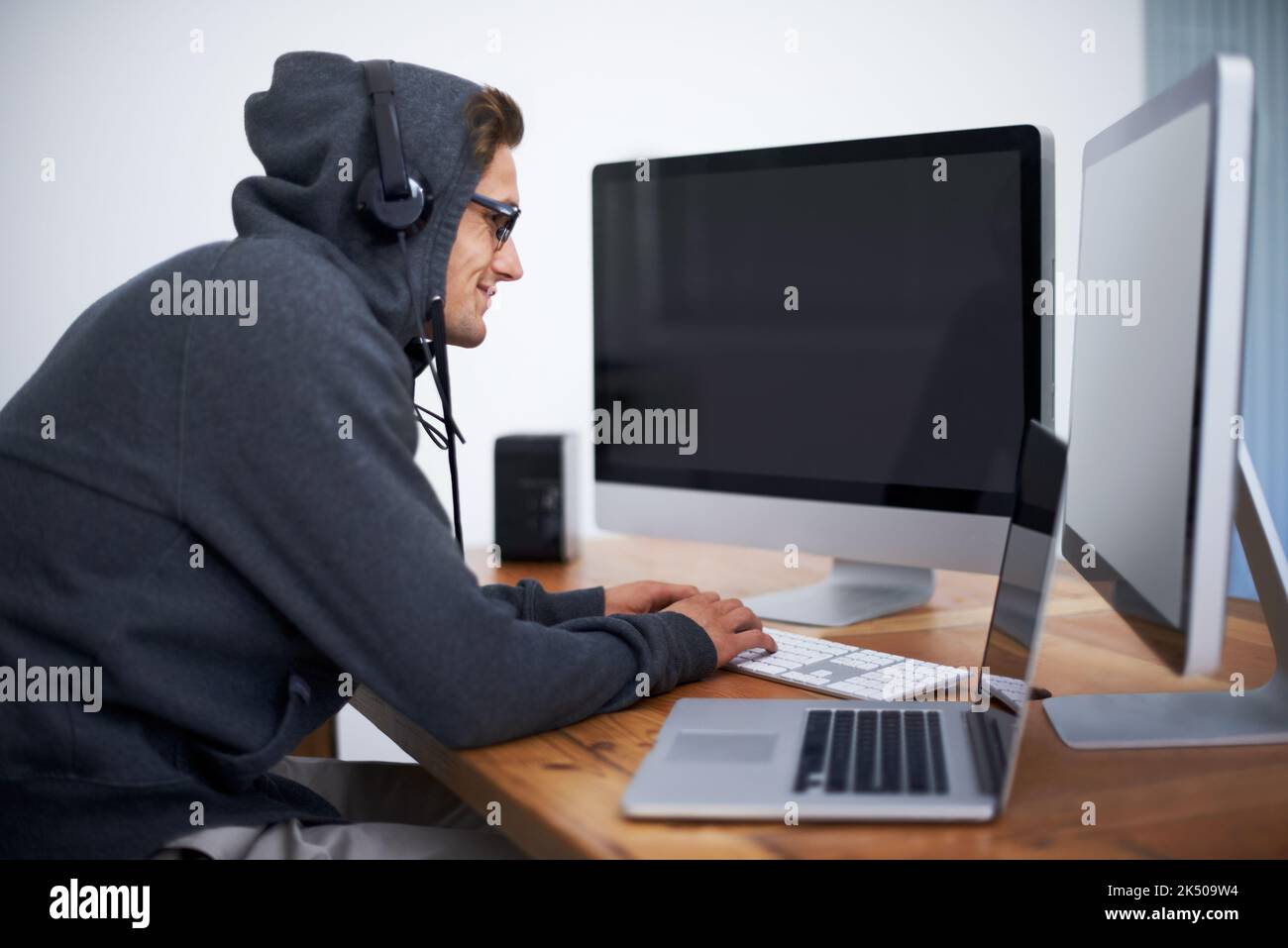 Nichts anderes als seine Arbeit ist in seinem Kopf. Ein junger Hacker, der auf zwei Bildschirmen und einem Laptop mit Hoodie arbeitet. Stockfoto