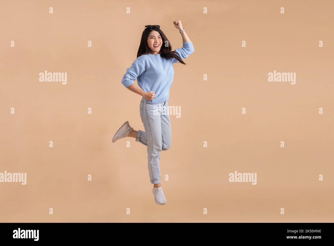 Glücklich lächelnde asiatische Frau mit Springen in der Luft hob ihre Hände in blauen Pullover und Jeans isoliert auf beigefarbenen Hintergrund. Ganzkörperlänge in Studioaufnahmen. Stockfoto