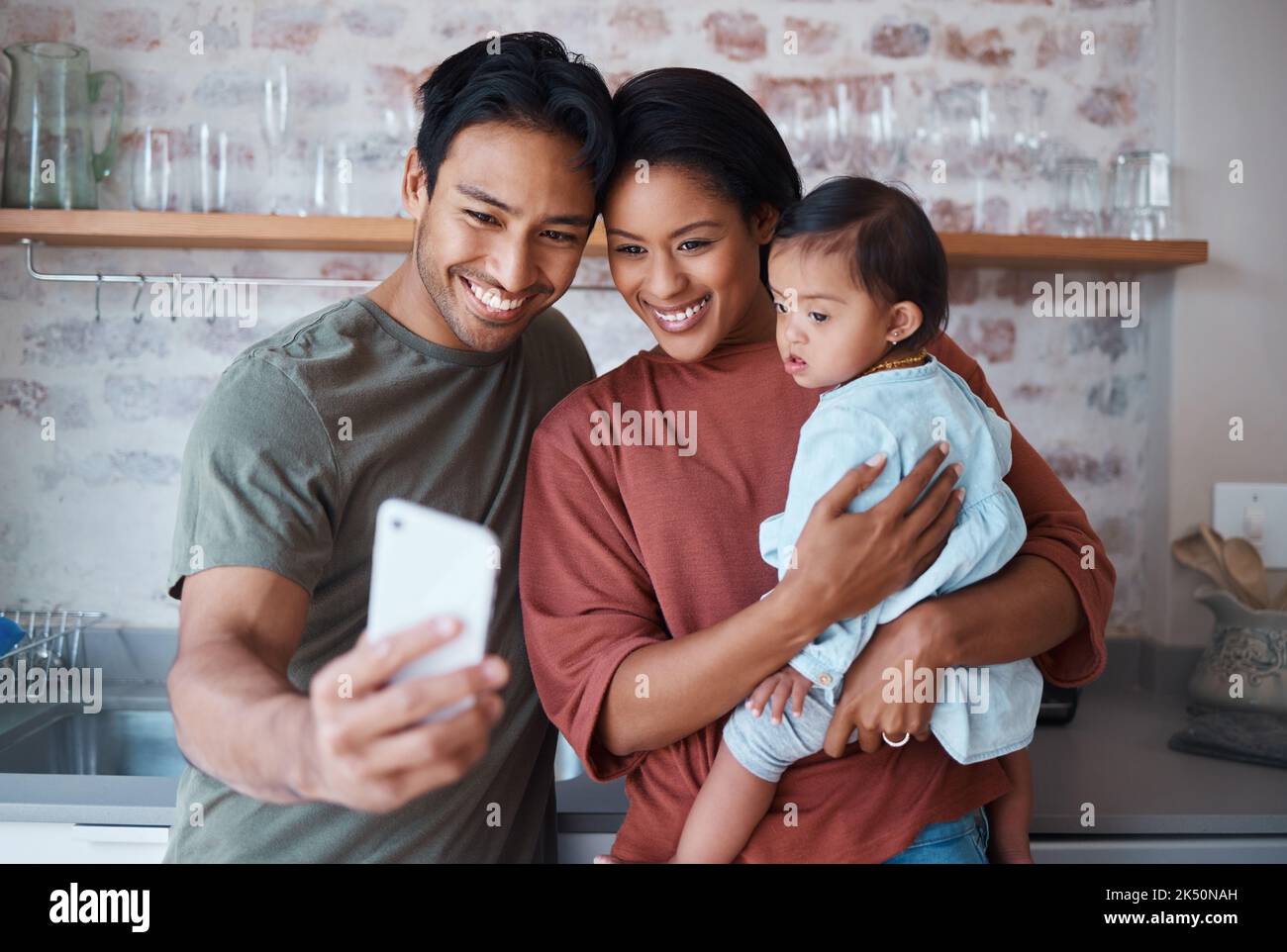 Glücklich, Eltern und Baby mit Down-Syndrom nehmen Selfie zusammen in der Küche des Familienhauses. Glück, Lächeln und Mutter und Vater verbinden sich mit Stockfoto