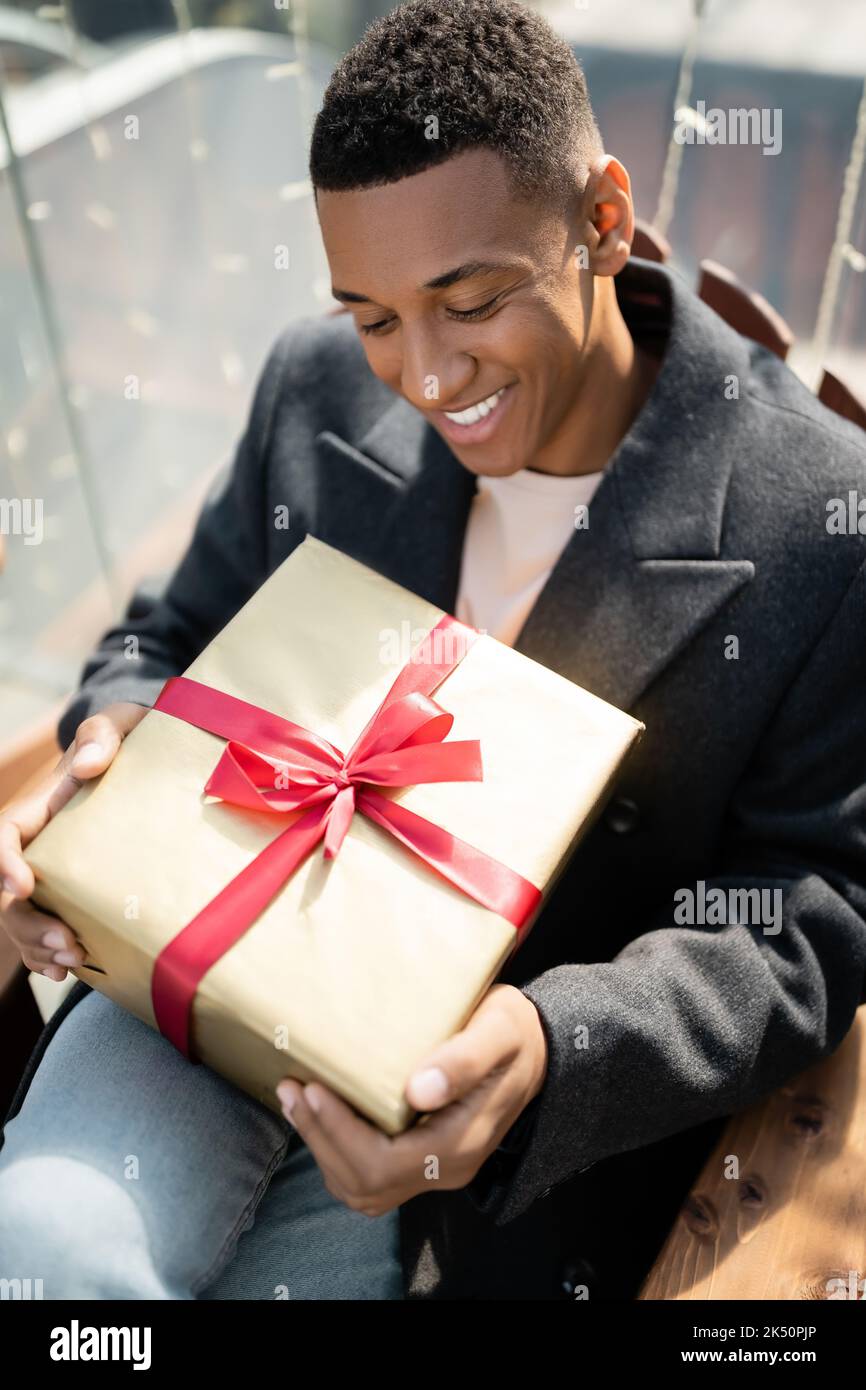Happy african american Mann im Mantel sitzt mit Geschenkbox im Freien, Stock Bild Stockfoto