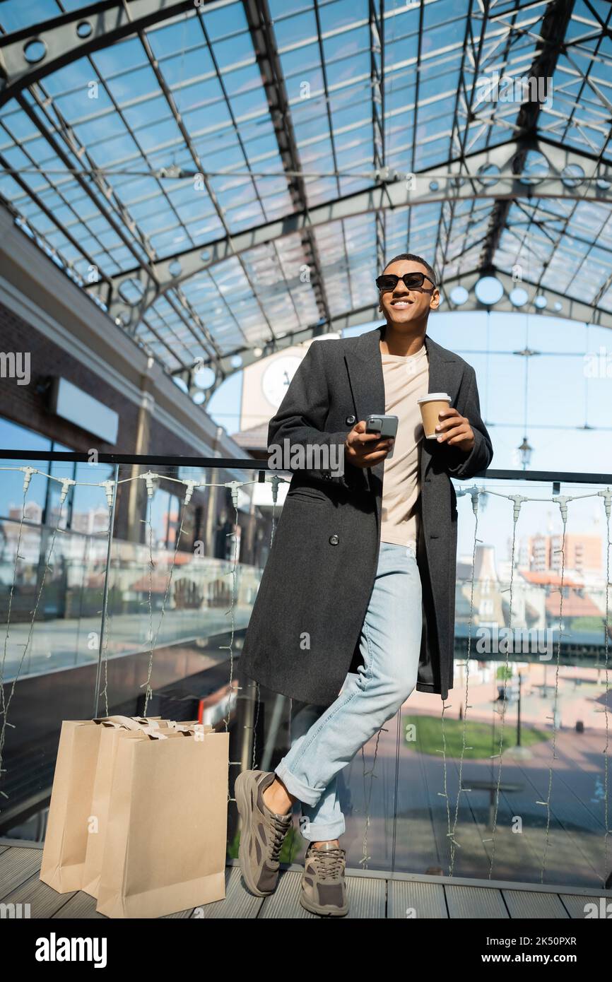 In voller Länge von afroamerikanischen Mann mit Kaffee zu gehen und Smartphone in der Nähe von Einkaufstaschen und Einkaufszentrum stehen, Stock Bild Stockfoto
