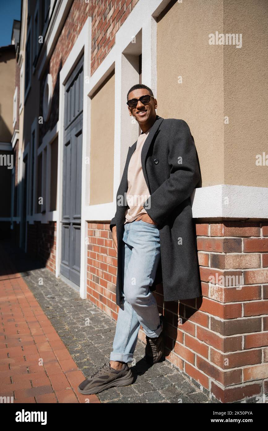 Ganzer afroamerikanischer Mann in Mantel und Sonnenbrille, der mit der Hand in der Tasche der Jeans in der Nähe des Gebäudes steht, Stockbild Stockfoto