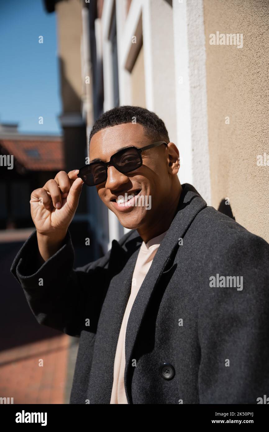 Trendiger afroamerikanischer Mann, der Sonnenbrille anpasst und die Kamera auf der Stadtstraße anlächelt, Stockbild Stockfoto