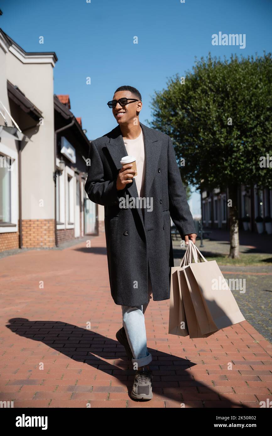 Ganze Länge des afroamerikanischen Mannes in trendigem Mantel und Sonnenbrille, der mit einem Getränk zum Mitnehmen und Einkaufstaschen spazierengeht, Stockbild Stockfoto