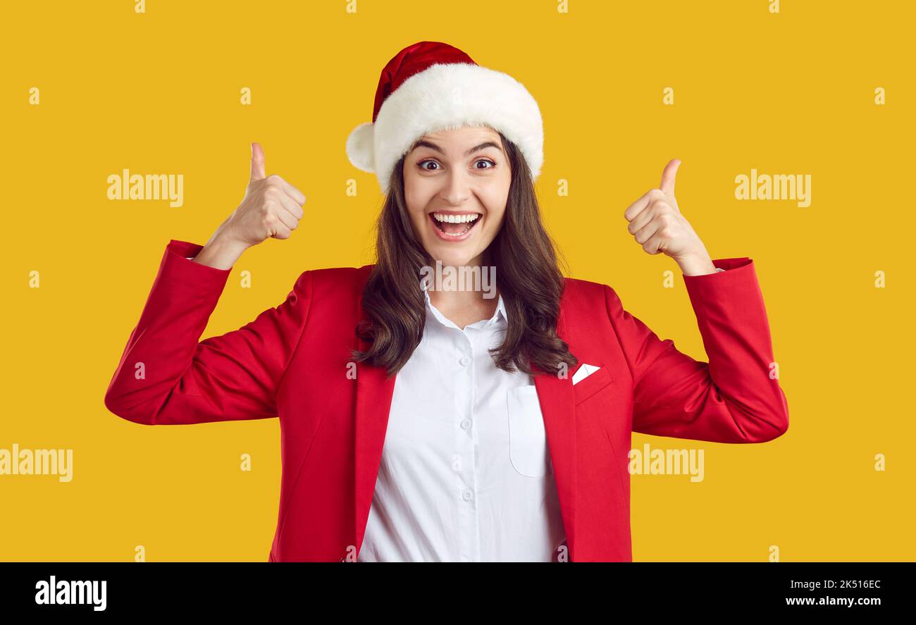 Fröhliche, aufgeregte junge Frau in rotem Anzug und Weihnachtshut, die Daumen hoch und lächelnd zeigt Stockfoto