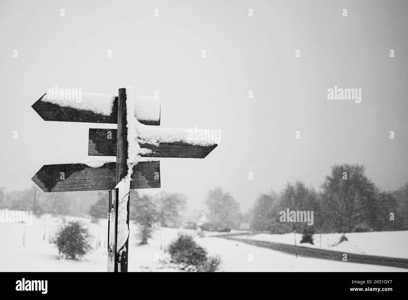 Schwarz und weiß von hölzernen Richtungs-Straßenschild auf schneebedecktem Feld gegen düsteren bewölkten Himmel in der Landschaft platziert Stockfoto