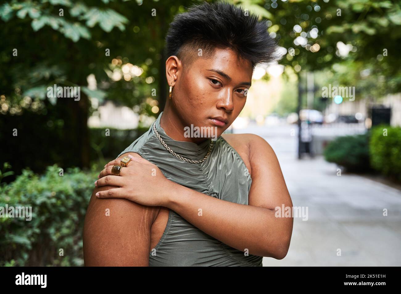 Asiatische Transgender-Frau mit kurzen Haaren und Pickel im Gesicht und Blick auf die Kamera, während sie auf unscharfem Hintergrund der Straße und des Parks in der Stadt steht Stockfoto