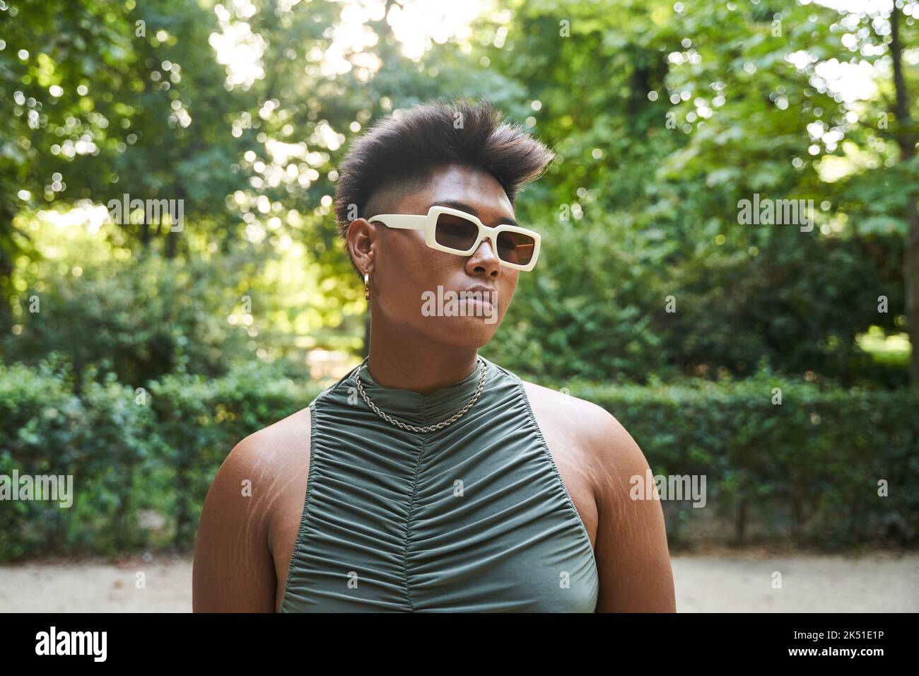 Philippinische Transgender-Person in stilvoller Sonnenbrille, die im Sommer auf den verschwommenen Hintergrund der Parkallee schaut Stockfoto
