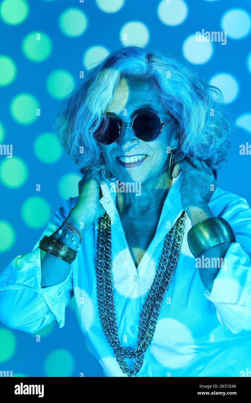 Glückliche ältere Frau mit stilvollen Accessoires lächeln und justieren Kragen des weißen Hemdes unter blauem Neonlicht und Projektion von Kreisen Stockfoto