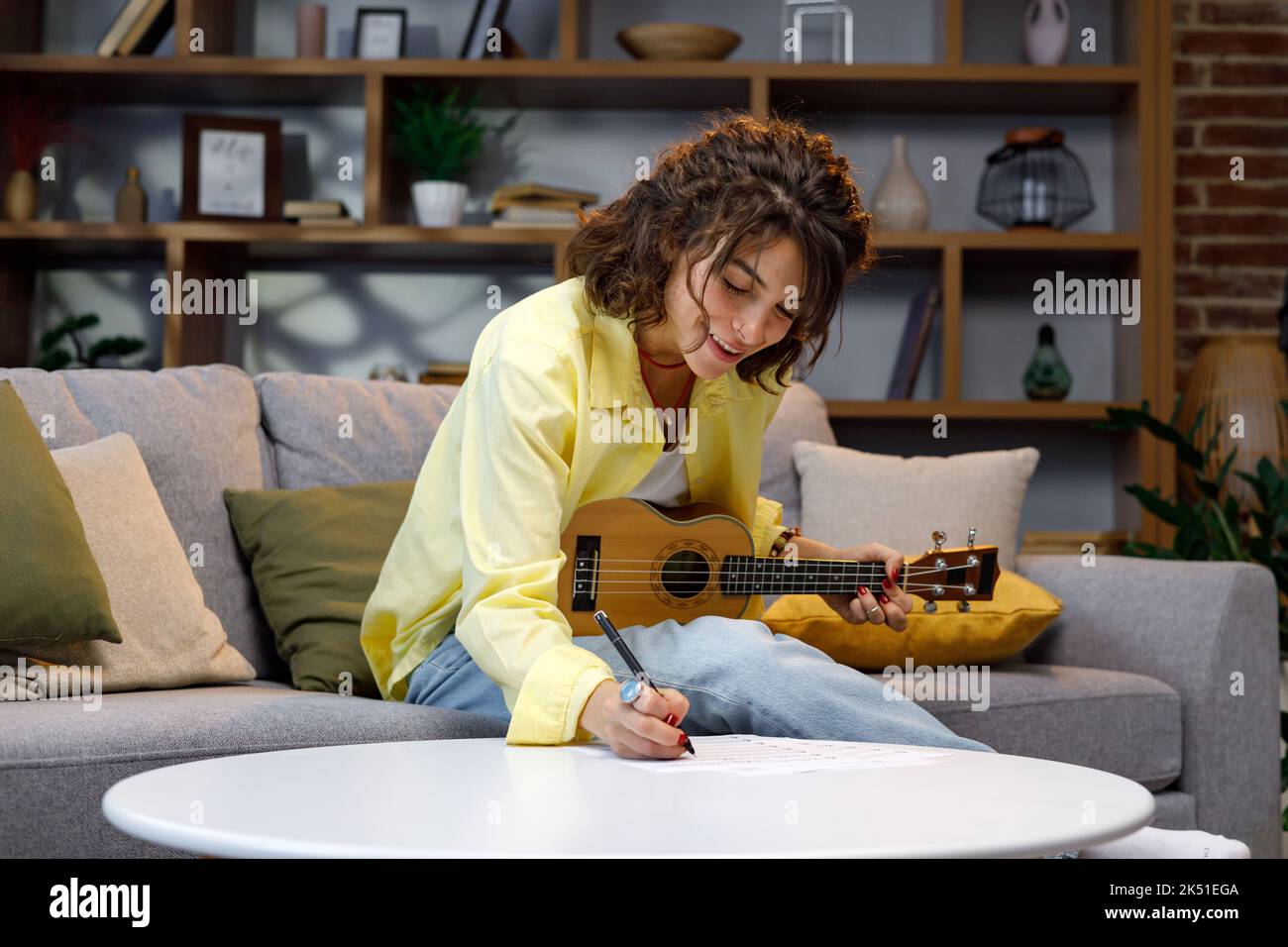 Ein schönes Hipster-Mädchen in einem gelben Hemd spielt und singt auf der Ukulele auf dem Sofa in der Wohnung. Komponiert ein Lied, schreibt Notizen. Startseite Hobbies Stockfoto