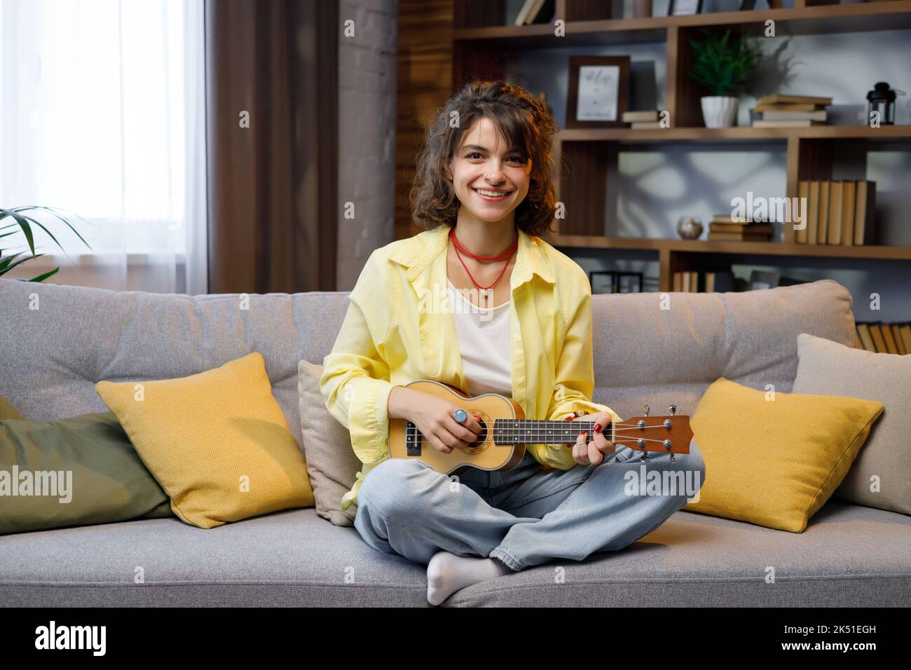 Ein schönes Hipster-Mädchen mit lockigen Haaren in einem gelben Hemd spielt und singt die Ukulele auf dem Sofa in der Wohnung. Startseite Hobbys spielen Musical Stockfoto
