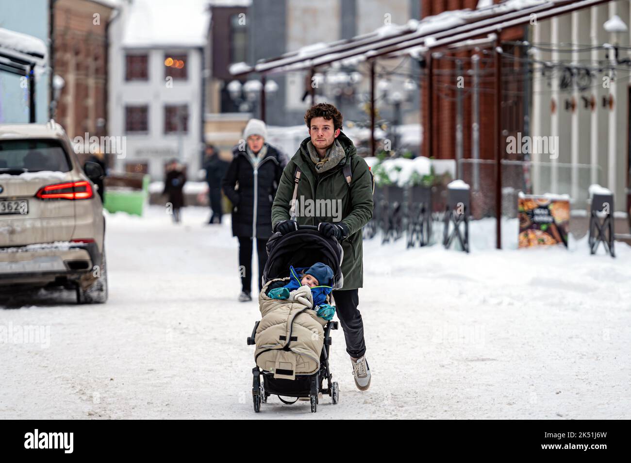 Riga, Lettland, 23. Dezember 2021: Junger erwachsener Vater schiebt Kinderwagen und läuft nach Schneesturm auf schneebedecktem Bürgersteig in der Stadt Stockfoto