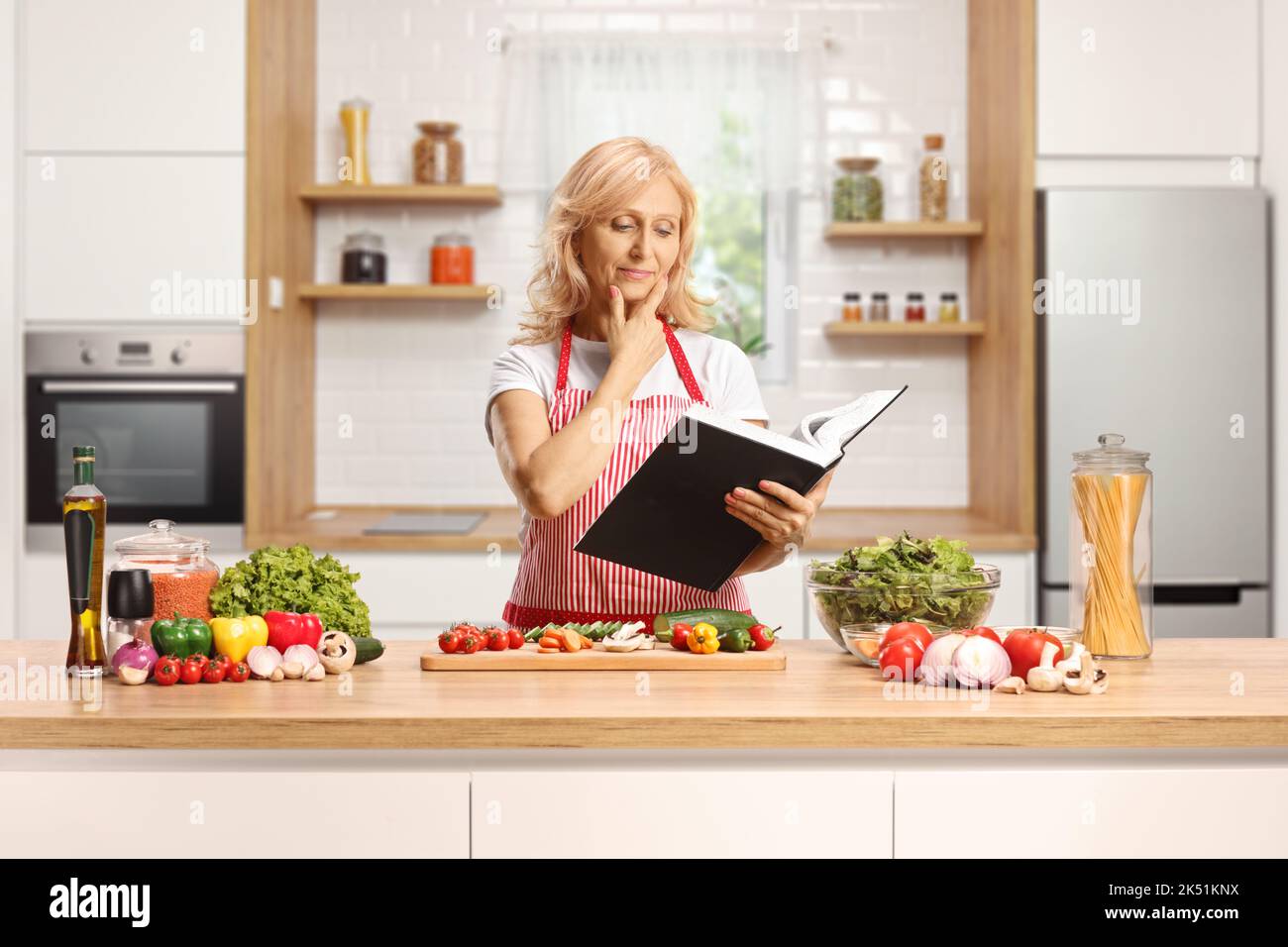 Hausfrau liest aus einem Kochbuch und steht hinter einer Küchentheke mit Gemüse Stockfoto